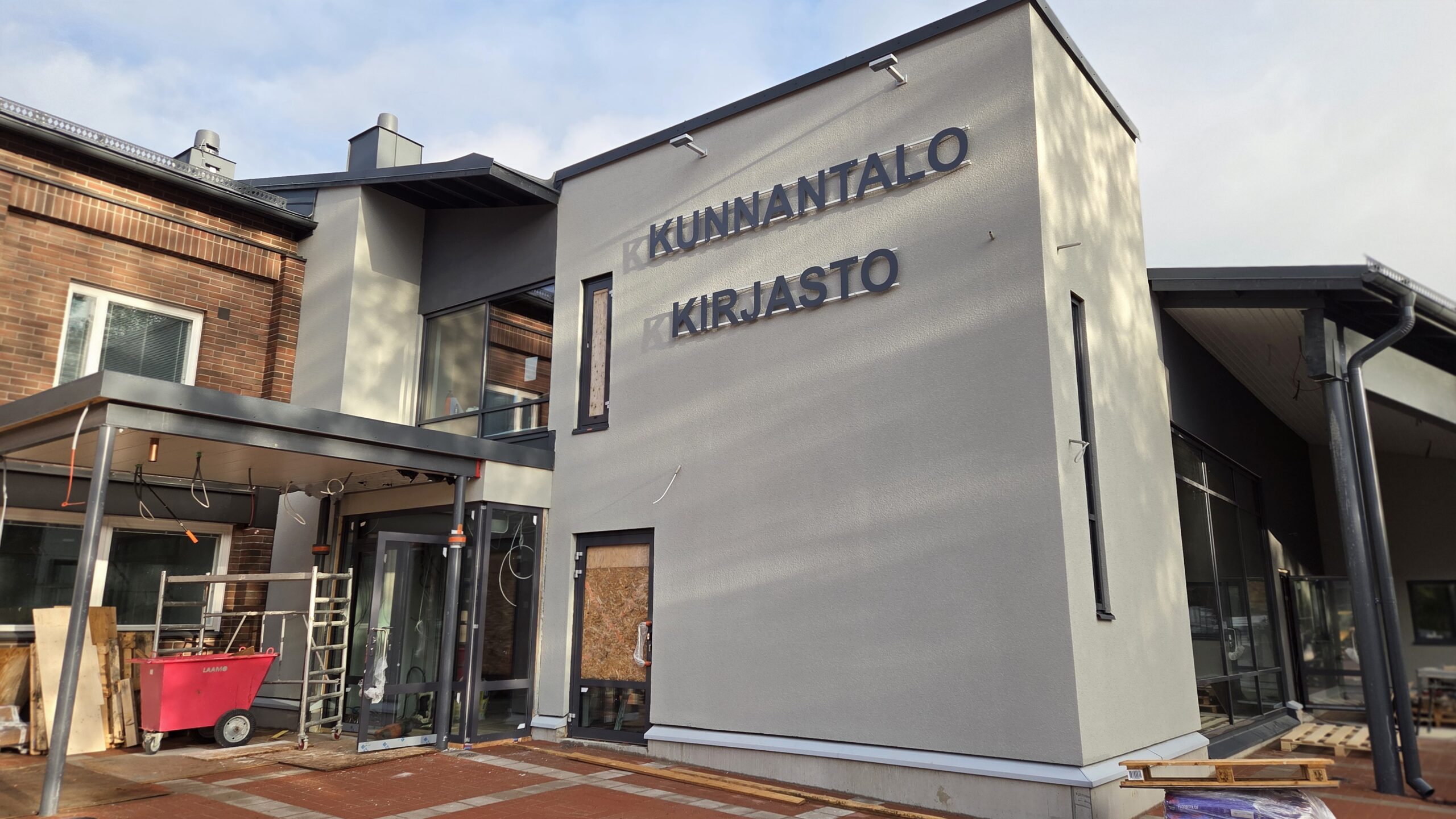 Ruskon kunnantalo pääovet, seinällä nimiö Kunnantalo Kirjasto. Rakennusmateriaaleja kivetyksellä.